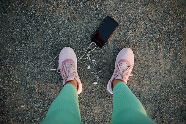 Vista dall'alto di uno smartphone nero con copia spazio pubblicitario per testo pubblicitario o auricolari per app mobili sdraiati su un campo sportivo vicino alle gambe dell'atleta femminile