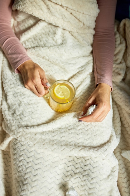 Vista dall'alto Donna malata che tiene in mano una tazza di tè caldo all'olivello spinoso e seduta sotto la coperta a casa, signora malata, rimedi naturali per il raffreddore, concetto di medicina naturale.