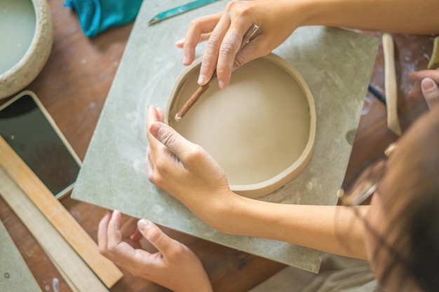 Vista dall'alto Donna vasaio che lavora al tornio del vasaio che fa vaso di ceramica dall'argilla nel laboratorio di ceramica Mano di messa a fuoco giovane donna che attacca la parte del prodotto in argilla al futuro prodotto in ceramica Laboratorio di ceramica