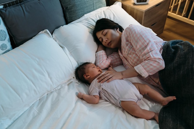 vista dall'alto la madre coreana stanca che indossa il pigiama è sdraiata addormentata sul letto con una mano appoggiata sull'innocente bambino che si sveglia in camera da letto durante l'ora di coricarsi la notte.