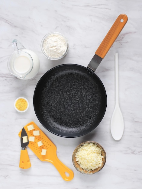 Vista dall'alto Padella, formaggio, farina, latte, preparazione di salsa besciamella (salsa bianca per pasta) Ingredienti sul tavolo di marmo bianco. Pentola Nera Vuota