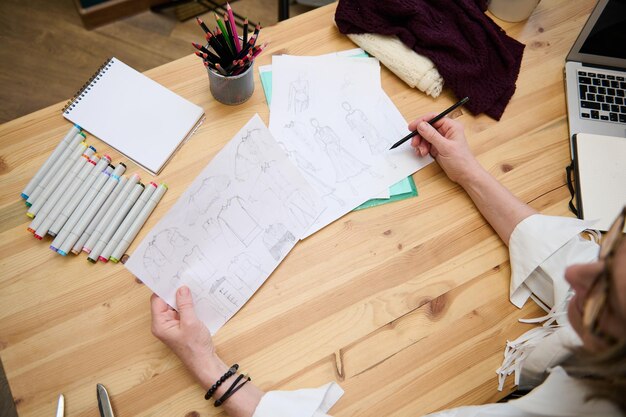 Vista dall'alto Primo piano di un anziano stilista di moda che lavora con schizzi per vestiti seduto alla scrivania di legno nel suo laboratorio creativo di design di moda