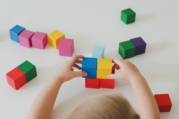 Vista dall'alto sulle mani del bambino che giocano e costruiscono con mattoni giocattolo di legno colorati su un tavolo di legno bianco e