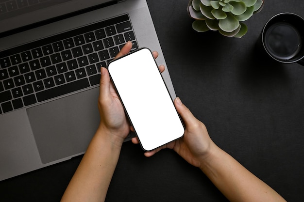 Vista dall'alto Un modello di smartphone è nelle mani di una donna sopra il computer portatile nella moderna scrivania da ufficio nera