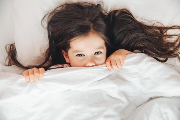 Vista dall'alto una bambina sdraiata a letto nascosta sotto una coperta che guarda la telecamera a casa