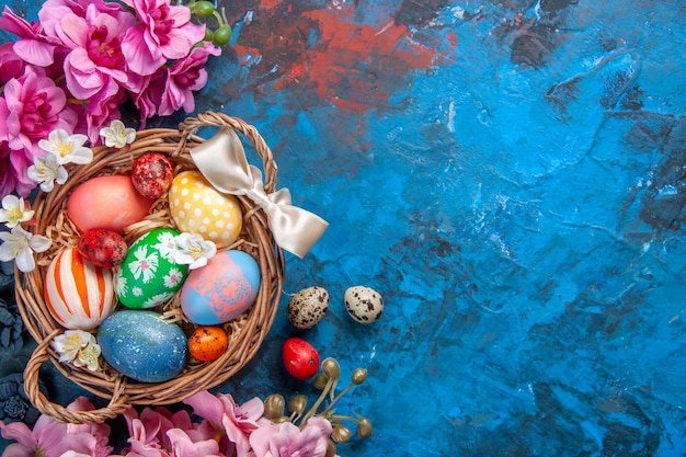 vista dall'alto uova di pasqua colorate all'interno del cesto con fiori su superficie blu