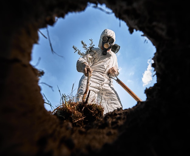 Vista dall'interno della fossa dell'ecologo che scava una fossa con la pala e piantando alberi indossando maschera antigas e tute protettive all'aperto sullo sfondo del cielo nuvoloso blu