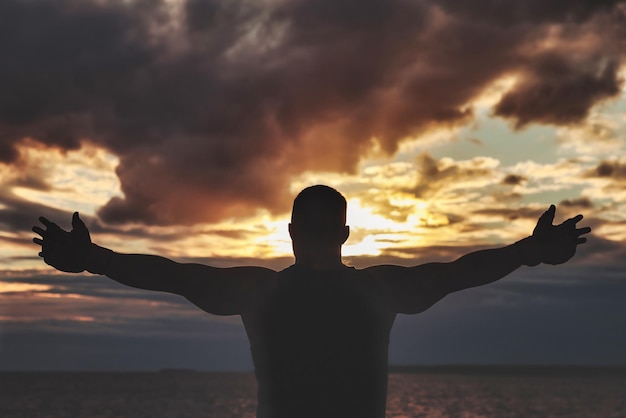 Vista dalla parte posteriore della silhouette di un uomo atletico con guanti da boxe in posa con le braccia alzate e bicipiti contro il cielo al tramonto