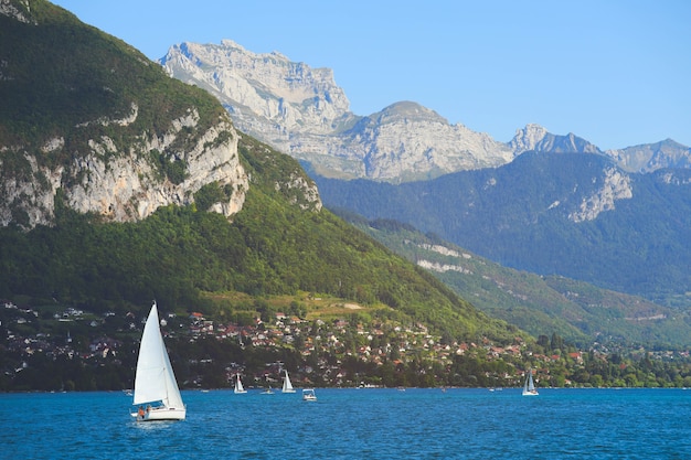 Vista del lago di Annecy con limpide acque turchesi, barche a vela e tradizionali case di legno in giornata estiva, France