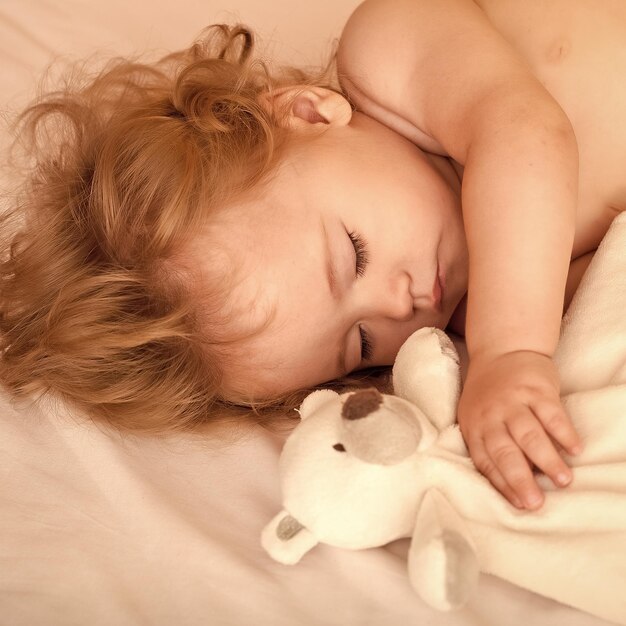 Vista del primo piano di adorabile bambino addormentato con capelli biondi ricci guance rotonde e dita minuscole sdraiato con gli occhi chiusi a letto con peluche orso ripieno giocattolo su sfondo bianco immagine quadrata