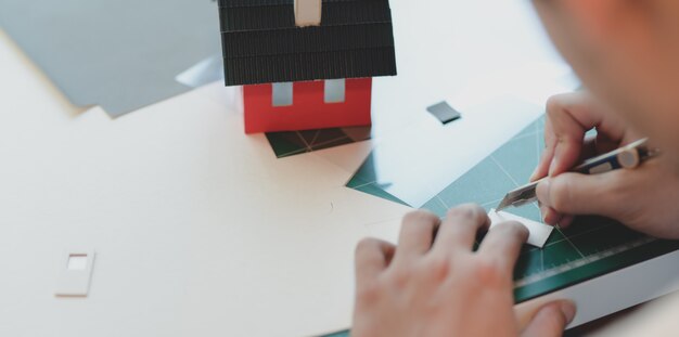 Vista del primo piano di giovane architetto maschio che fa il modello della casetta mentre tagliando il cartone