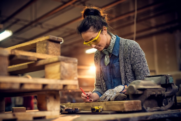 Vista del ritratto del lavoratore di carpenteria femminile professionale invecchiato mezzo sorridente soddisfatto con un disegno a matita sulla misura di nastro e di legno, morsa d'acciaio sulla tavola nell'officina.