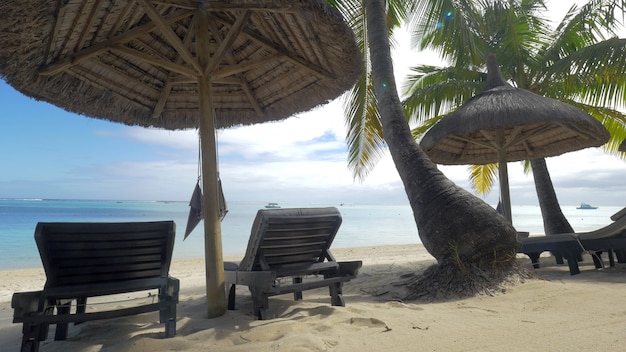 Vista della chaiselongue vuota vicino all'ombrellone nativo e alle palme contro l'isola mauritius dell'acqua blu