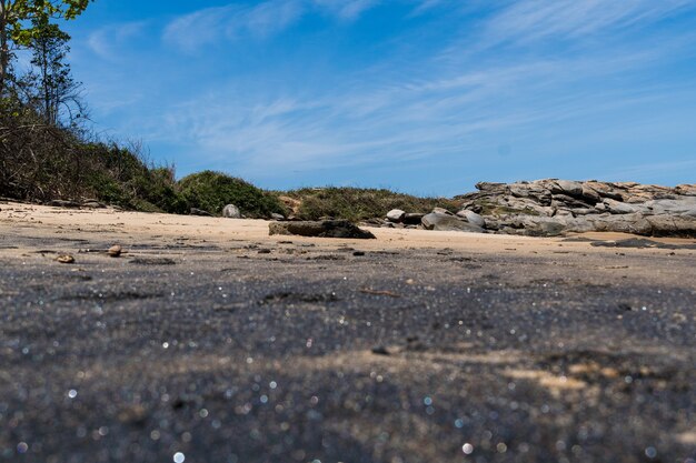 Vista della spiaggia di Areia Negras a Rio das Ostras a Rio de Janeiro con giornata di sole, cielo blu e alcune nuvole. Mare forte e sabbia nera mista a gialla e tanti scogli.