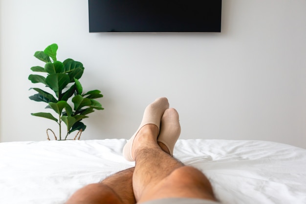 Vista delle gambe dell'uomo sul letto bianco con TV e pianta. Concetto di relax.