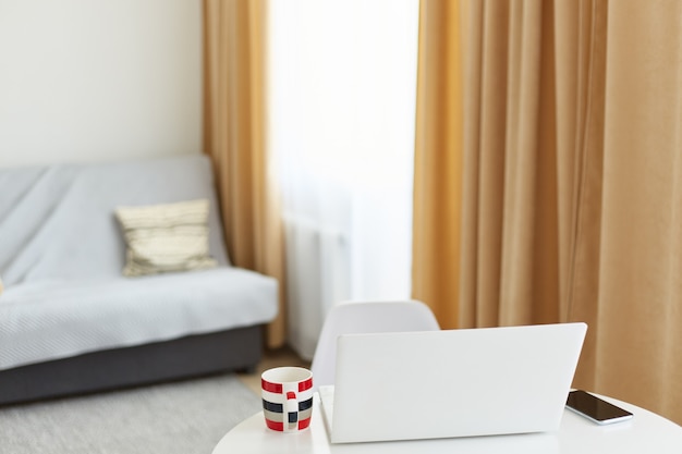 Vista dettagliata interna di un computer portatile bianco a casa in soggiorno con caffè o tè in tazza sul tavolo, area di lavoro vicino a una finestra con tende.