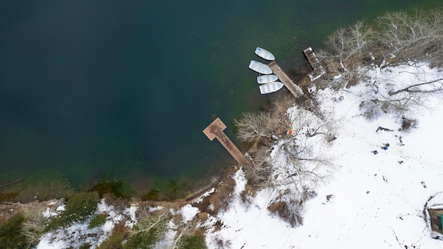 Vista drone del molo con barche, alberi, acqua limpida e liscia, neve sdraiata a terra.
