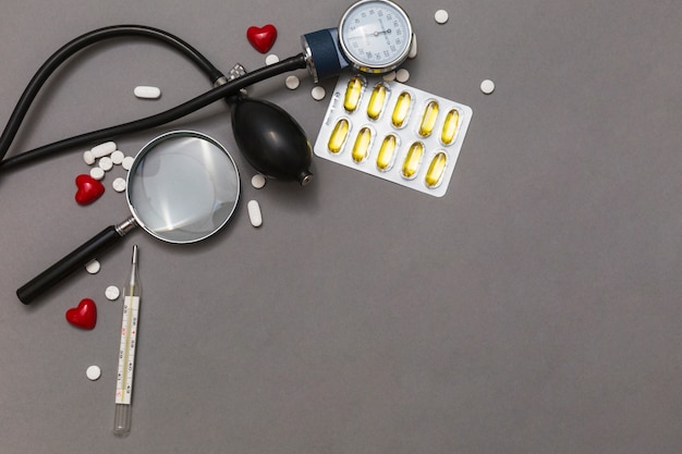 Vista elevata dello sfigmomanometro; lente d&#39;ingrandimento; pillole; termometro e cuore rosso su sfondo grigio