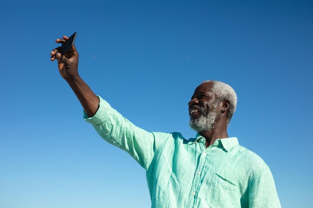 Vista frontale ad alto angolo di un anziano uomo afroamericano in piedi sulla spiaggia con cielo blu sullo sfondo, sorridente e prendendo selfie