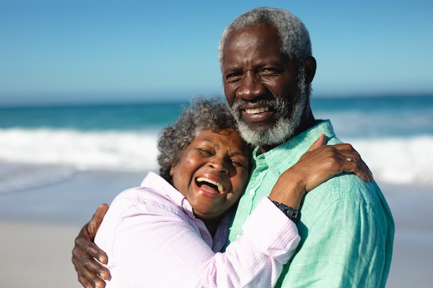 Vista frontale da vicino di una coppia afroamericana anziana in piedi sulla spiaggia con cielo blu e mare sullo sfondo, abbracciando, ridendo e sorridendo