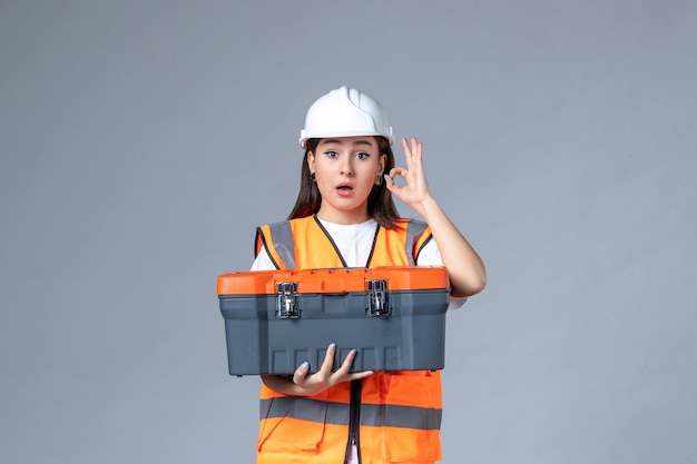 Vista frontale del costruttore femminile che tiene la cassetta degli attrezzi sul muro grigio