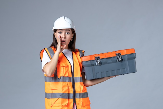 Vista frontale del costruttore femminile che tiene la cassetta degli attrezzi sul muro grigio