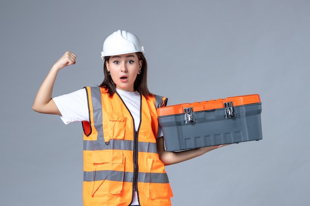 Vista frontale del costruttore femminile che tiene la cassetta degli attrezzi sul muro grigio