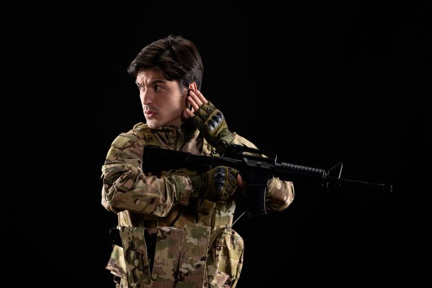 Vista frontale del giovane soldato in uniforme con il fucile sul muro nero