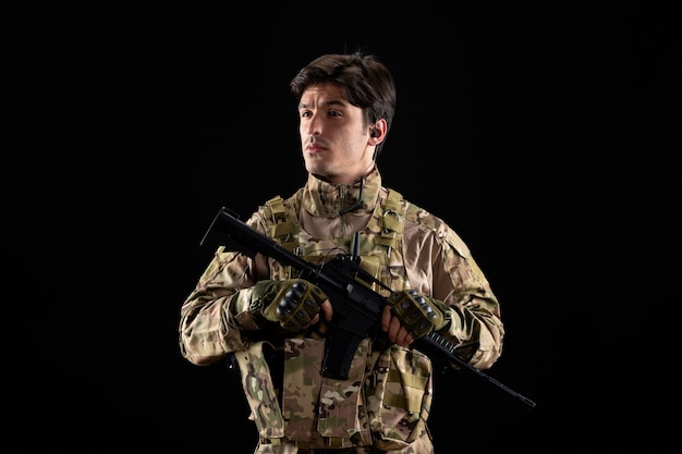 Vista frontale del giovane soldato in uniforme con il fucile sul muro nero