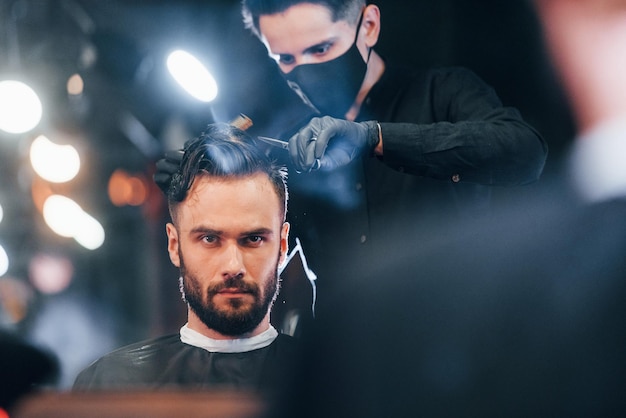 Vista frontale del giovane uomo barbuto che si siede e si fa tagliare i capelli nel negozio di barbiere da un ragazzo con una maschera protettiva nera