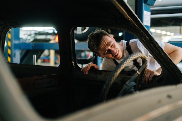 Vista frontale del giovane uomo di servizio frustrato in uniforme che ispeziona l'interno della vecchia auto in riparazione auto ...