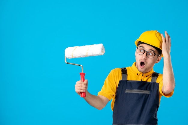 Vista frontale del lavoratore di sesso maschile in uniforme e casco con rullo di vernice su un blu