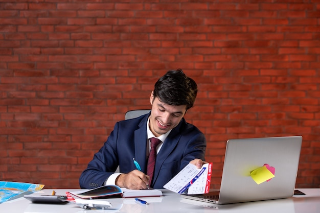 vista frontale del lavoratore di sesso maschile seduto dietro il suo posto di lavoro in tuta con in mano un paio di biglietti aerei piano di lavoro occupazione aziendale progetto estero lavoro