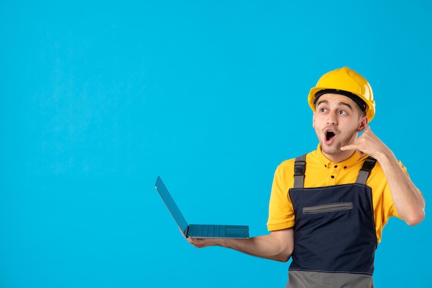 Vista frontale del lavoratore maschio in uniforme che lavora con il computer portatile e che posa sull'azzurro