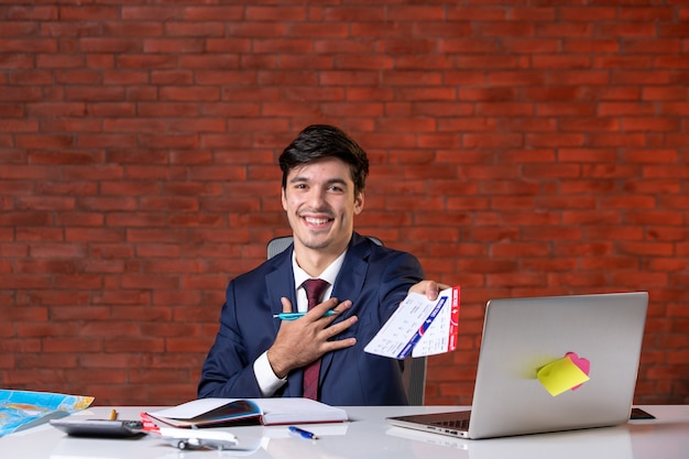 vista frontale del lavoratore maschio sorridente seduto dietro il suo posto di lavoro in tuta con in mano un paio di biglietti aerei piano di lavoro lavoro occupazione turismo aziendale progetto estero