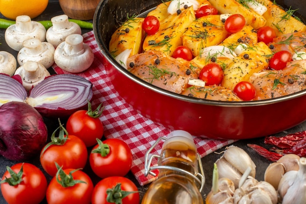 Vista frontale del pasto di pollo crudo con patate verdure in casseruola su asciugamano rosso spogliato funghi verdure fresche olio caduto bottiglia su sfondo grigio