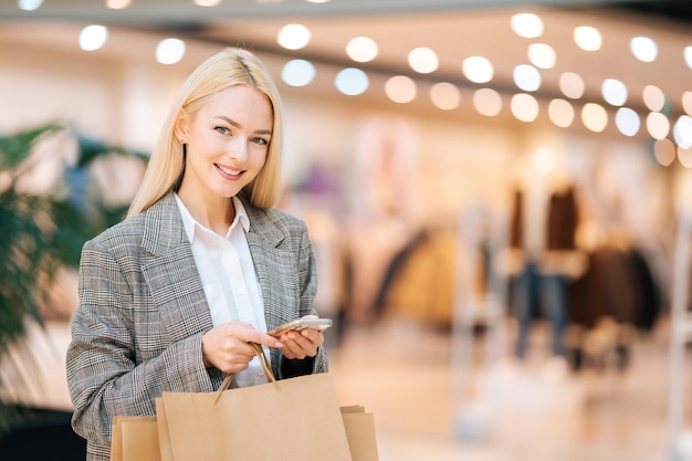 Vista frontale dell'eleganza sorridente giovane donna bionda in abiti eleganti guardando la fotocamera in possesso di telefono cellulare e sacchetti di carta per la spesa con gli acquisti nel centro commerciale, sfondo sfocato.