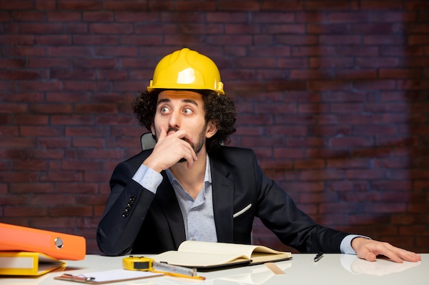 vista frontale dell'ingegnere maschio seduto dietro il posto di lavoro in tuta e casco giallo piano business builder occupazione lavoro appaltatore progetto di lavoro