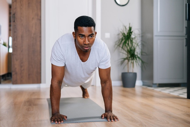 Vista frontale dell'uomo afroamericano concentrato che fa pushup sul pavimento nella stanza domestica che guarda lontano