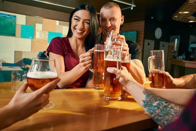 Vista frontale della coppia allegra seduta al tavolo al bar e ridendo con gli amici mentre si beve birra. Giovani che riposano nei fine settimana in compagnia in pub. Concetto di tempo libero e felicità.