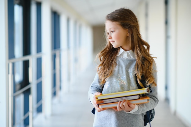 Vista frontale della piccola bella ragazza della scuola tra il corridoio della scuola che tiene le note a portata di mano Ragazza divertente e felice che sorride alla macchina fotografica che riposa dopo le lezioni della scuola primaria