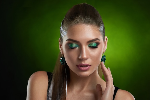 Vista frontale della sensuale ragazza bruna con trucco verde lucido che tocca la pelle bronzo perfetta del viso. Bella donna con gli occhi chiusi, indossa in top nero, grande orecchino arrotondato in posa seducente.