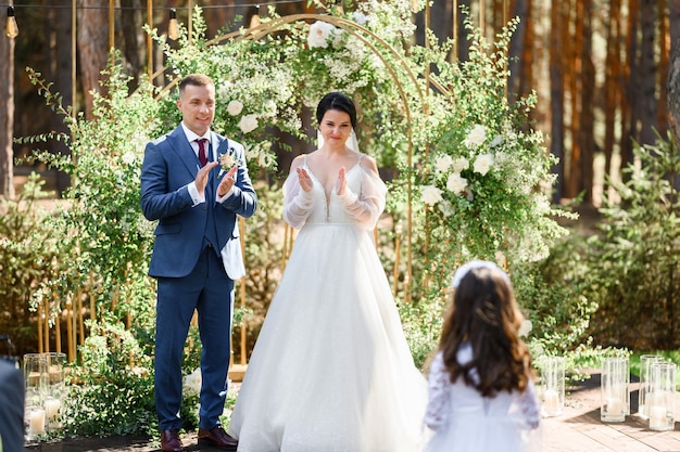 Vista frontale della sposa e dello sposo felici in abbigliamento festivo insieme in piedi sullo sfondo dell'arco nuziale decorato