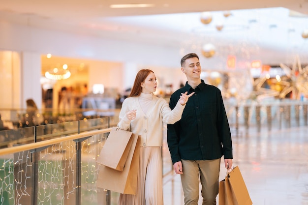 Vista frontale di belle giovani coppie felici che tengono borse della spesa che camminano nel centro commerciale, sfondo sfocato. Uomo bello gioioso e donna attraente che acquistano insieme al centro del negozio