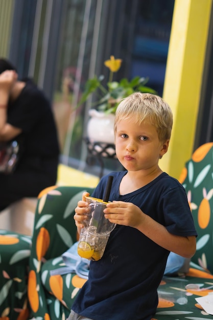 Vista frontale di felice ragazzo biondo assetato che beve limonata tè alla frutta kombucha tè freddo con pesca e limone benessere aranciate bevande dal bicchiere di plastica con paglia in piedi Storie formato verticale