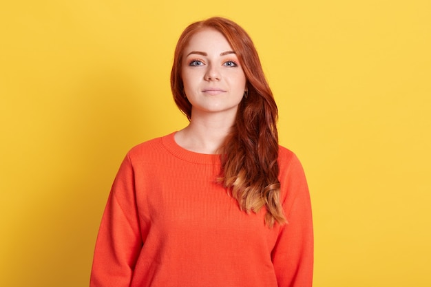 Vista frontale di grave giovane donna attraente con i capelli rossi che indossa un maglione arancione casual