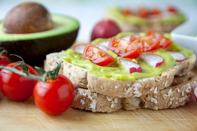 Vista frontale di pane tostato con avocado, pomodoro e ravanello e gli ingredienti sul tavolo di legno