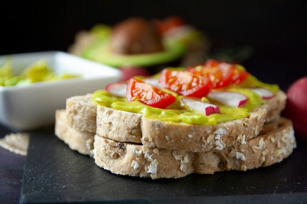 Vista frontale di pane tostato con avocado, pomodoro e ravanello e gli ingredienti sul tavolo nero