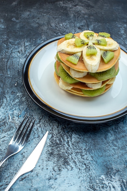 Vista frontale di soffici pancake in stile americano realizzati con yogurt naturale e impilati con strati di frutta su piatto e posate su sfondo di ghiaccio con spazio libero