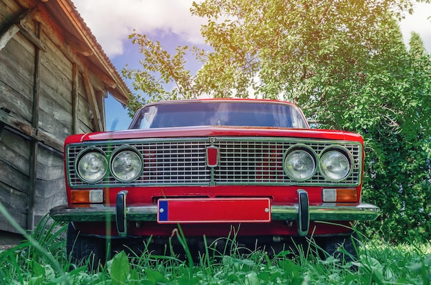 Vista frontale di un'auto Lada rossa d'epoca in campagna Vecchio fienile in legno dal lato Vecchia macchina rossa sovietica